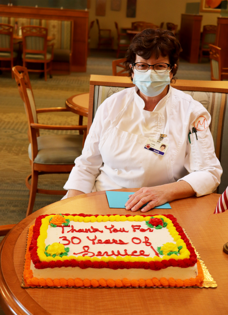 a woman in a chef's coat sitting in a booth behind a cake that reads "Thank you for 30 Years of Service" on a table 