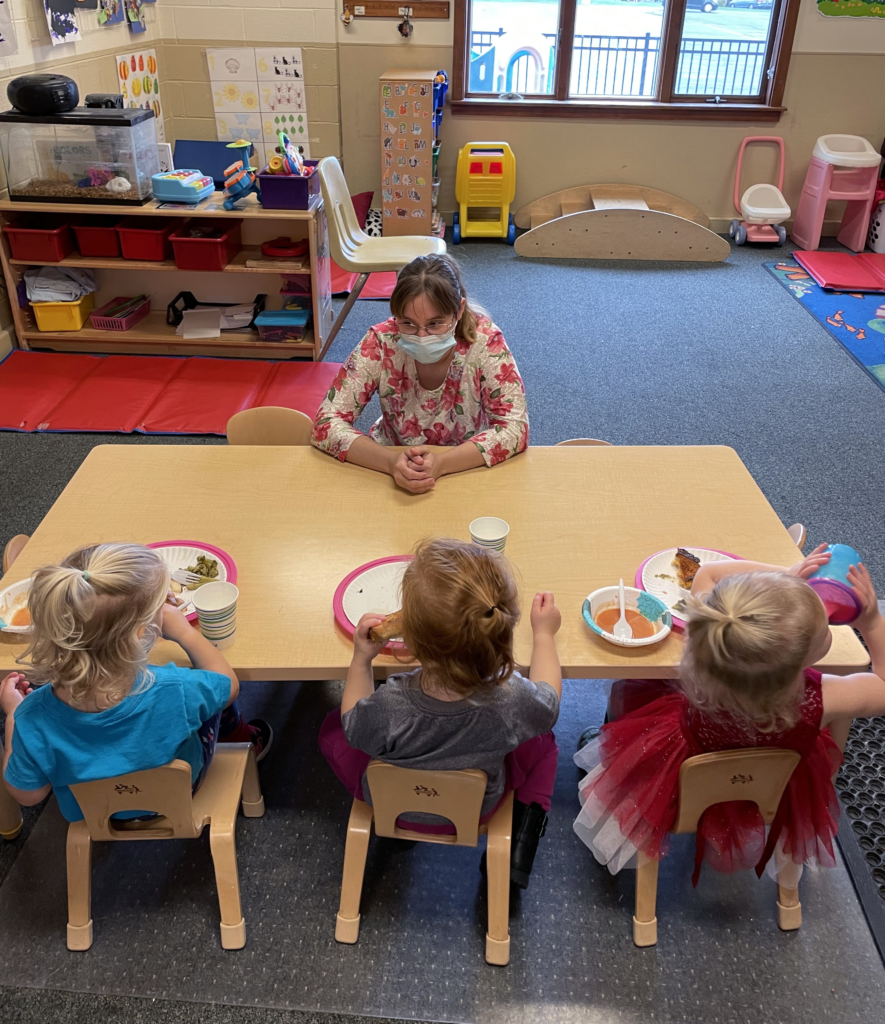 A teacher attending to three young students during a meal in a childcare center.