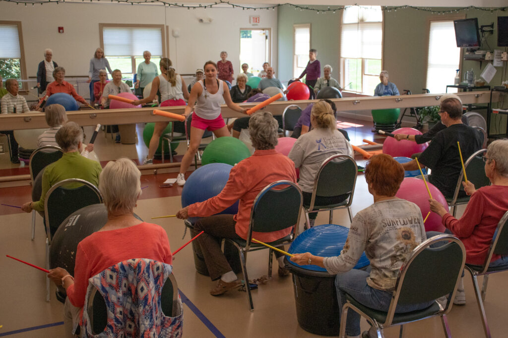 Emily Robarge standing at the front of a room with pool noodles in hand, drumming on a fitness ball. Seveal residents in chairs do the same in front of her.