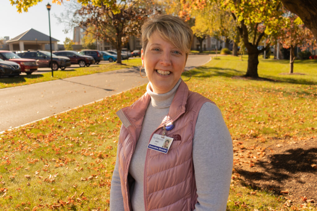 Nikki Rineer from the waist up standing with a smile on a lawn. There are trees and cars behind her.